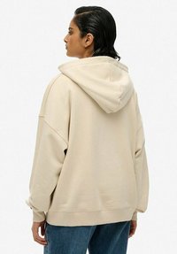 Haut à capuche beige oversize avec cordon de serrage, manches côtelées et ourlet. Tissu doux montré de côté et de dos, associé à un jean bleu.