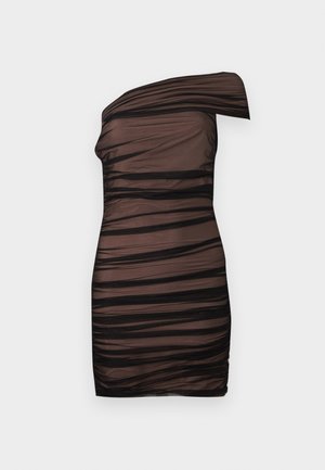 Femme debout portant une mini-robe noire asymétrique en voile froncé et des talons hauts beiges ouverts devant sur un fond uni.