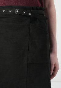 Sorte semsket forkle-lignende skjørt med belte i midjen, detaljer med øsken, sidelomme og en glatt, matte tekstur.