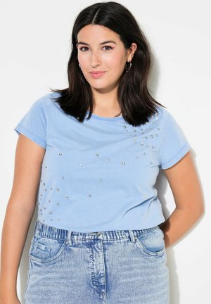 Lysblå kortærmet t-shirt med en blød tekstur, udsmykket med metal dekorative nitter spredt over forsiden. Sammen med denimshorts.