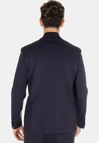 Uomo con capelli ricci corti che indossa un giacca scura di colore navy, visto di spalle su uno sfondo bianco semplice.