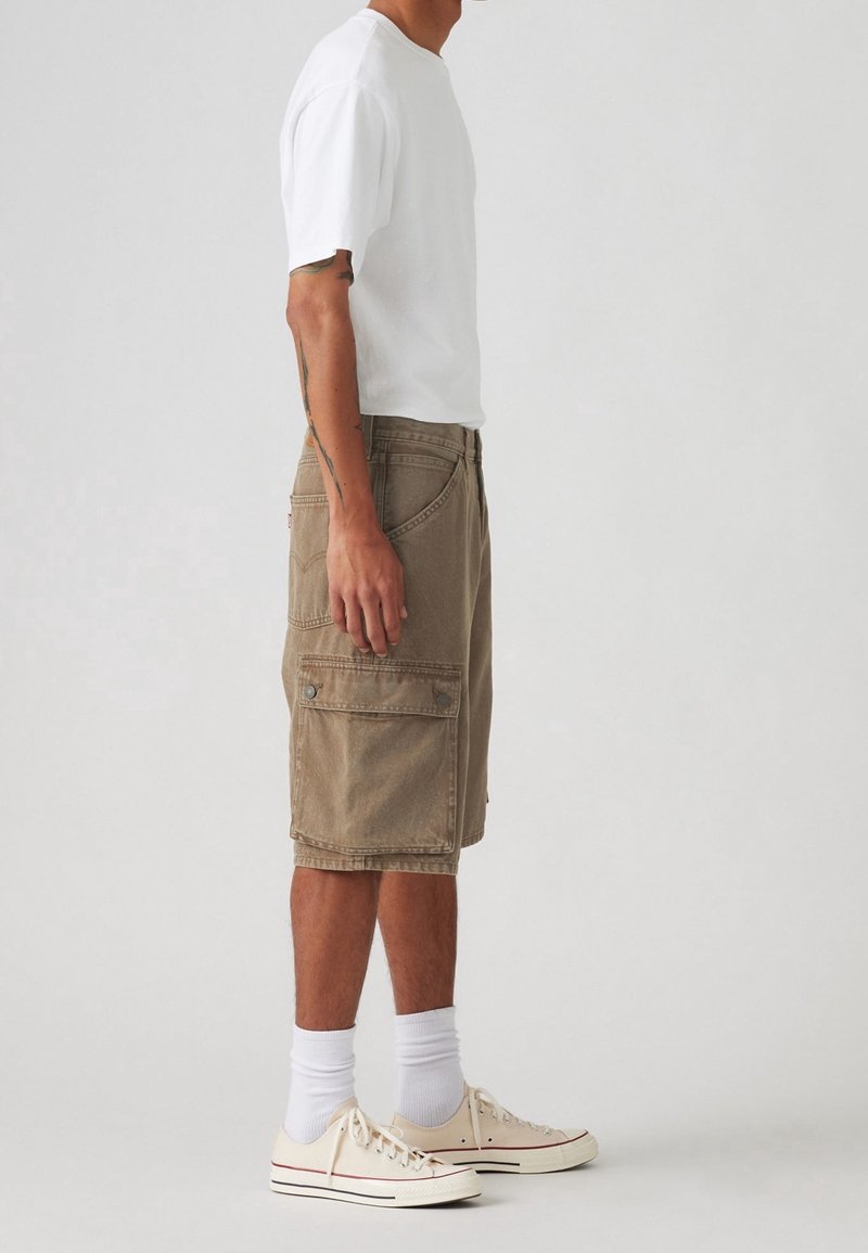 Beige cargo shorts made of cotton feature side pockets and a relaxed fit. Paired with a white t-shirt and beige sneakers with white socks.