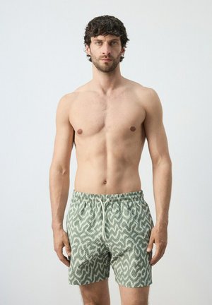 Hombre sin camiseta con cabello rizado oscuro que lleva puestos unos pantalones cortos de baño verdes con estampado, de pie contra un fondo claro y liso.