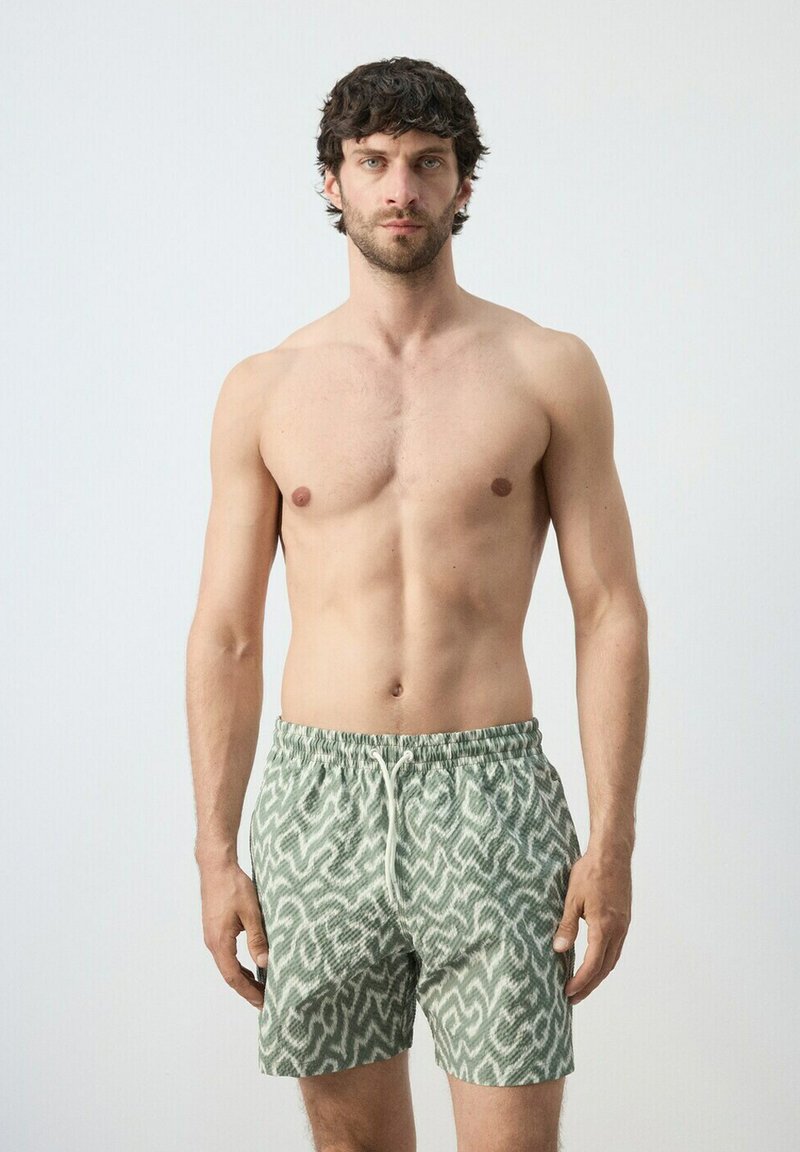 Homme torse nu aux cheveux bouclés foncés portant un short de bain vert à motifs, debout devant un fond clair uni.