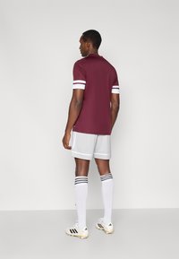 Bordeauxrotes Polohemd mit weißen Streifen an den Ärmeln, kombiniert mit grauen Shorts und weißen Socken mit schwarzen Streifen. Die Fußballschuhe sind cremefarben mit schwarzen Akzenten.