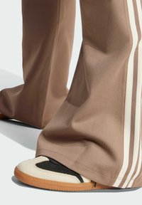 Bruna, löst sittande joggers med vita sidosträck, kombinerat med svart- och vitfärgade sneakers med en gummisåle. Fokus på skoddets detaljer.