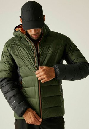 Mężczyzna w czarnej czapce zapina zieloną i czarną pikowaną kurtkę z kapturem na jednolitym tle.