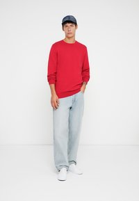 Pull en maille rouge avec un col rond, manches longues, associé à un jean baggy bleu clair et des baskets blanches. Une casquette bleue complète le look.