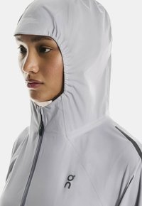 Svetlo siva jakna s kapuco z gladko, mat površino. Ima zadrgo spredaj in črne logotipne akcentne detajle. Oprijet kroj z nabranimi deli kapuce.