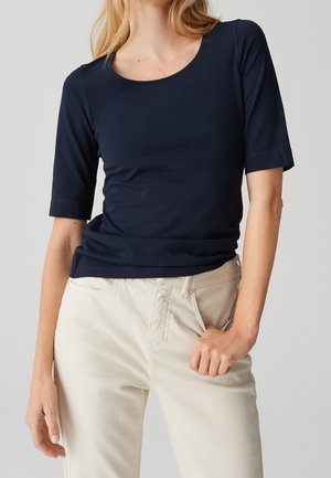 Woman wearing a fitted navy blue half-sleeve shirt and off-white pants, with one hand in pocket against a plain background.