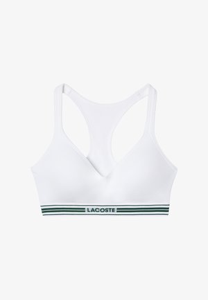 Top sportivo bianco con scollatura a V, caratterizzato da un bordo striato verde nella parte inferiore con il testo "LACOSTE". Tessuto liscio e stile a corsetto.