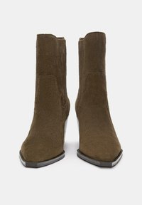 Une paire de bottines en daim marron avec des orteils pointus, des talons bas empilés et des panneaux latéraux élastiques, présentée de face sur un fond blanc.