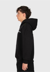Junger Mann mit lockigem Haar, der einen schwarzen Hoodie mit weißem Text trägt und im Profil vor einem schlichten hellen Hintergrund steht.