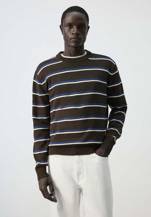 Hombre con suéter de rayas marrón, azul y blanco y pantalones blancos, de pie con una mano en el bolsillo frente a un fondo liso.