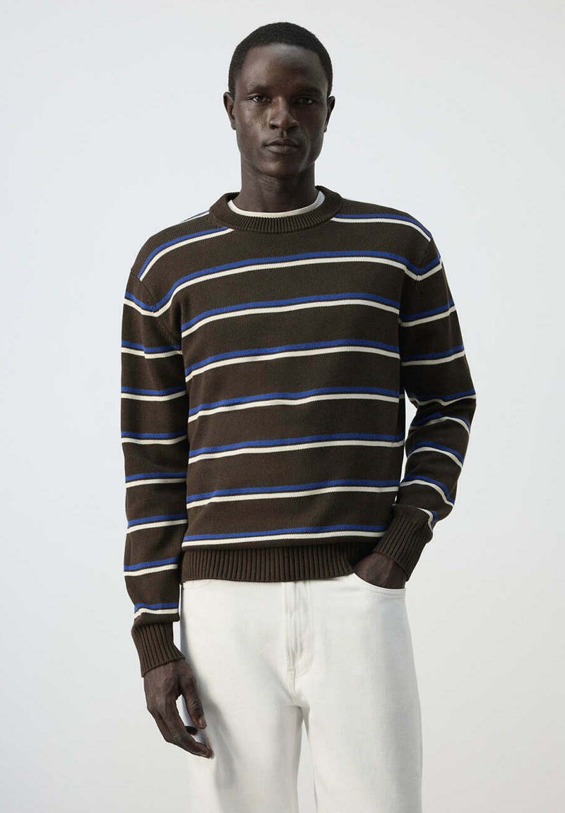 Homme portant un pull rayé marron, bleu et blanc et un pantalon blanc, debout avec une main dans la poche sur un fond uni.