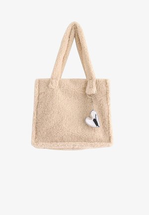 Borsa tote in finta pelliccia beige con due manici in tessuto, forma quadrata e un charm a forma di cuore in argento attaccato al lato.