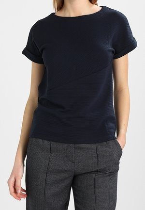 Femme portant un haut texturé bleu marine à manches courtes et un pantalon gris sur mesure avec poches, debout avec une main dans la poche.