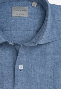 Jasnoniebieska koszula w jodełkę z kołnierzykiem typu fold-down, pojedynczym białym guzikiem przy szyi oraz szarym metkiem z informacją o rozmiarze i marce.