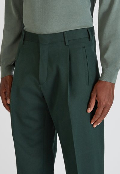 Dunkelgrüne Hose aus strukturiertem Stoff, mit flachem Frontdesign, seitlichen Taschen und einer taillierten Passform. Kombiniert mit einem hellgrünen Oberteil.