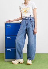 Vit kortärmad t-shirt med gul Adidas-logotyp, tillsammans med ljusblå vida jeans. Modellen står bredvid en blå arkivskåp.