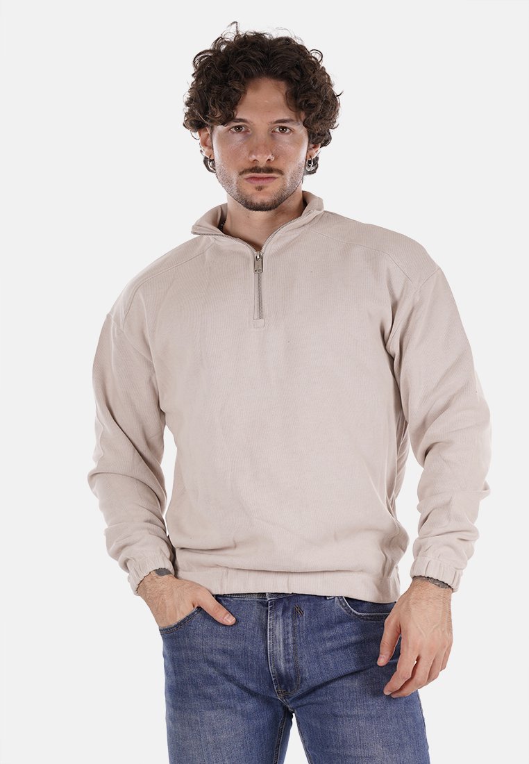 Felpa beige con zip, texture a coste, maniche lunghe, polsini aderenti e colletto alto. Indossata con jeans blu scuro, in una posa neutra.