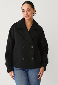 Schwarzer doppelt gebundener Peacoat mit großen Revers, Taschen und Knopfakzenten, aus einem strukturierten Stoff gefertigt, über hellblauen Jeans getragen.