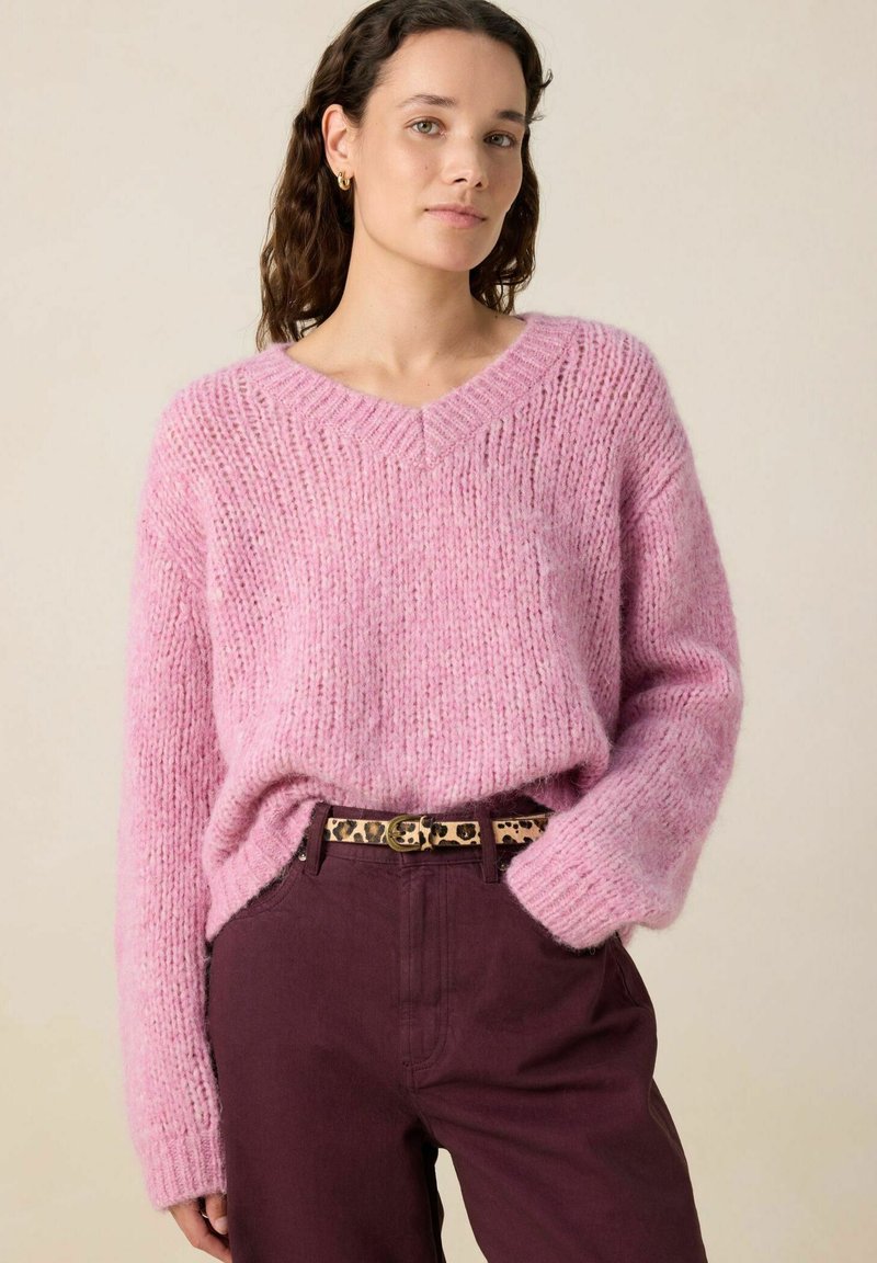 Pull en tricot rose avec un col en V, présentant une coupe ample et une texture duveteuse, associé à un pantalon bordeaux et une ceinture imprimée léopard.