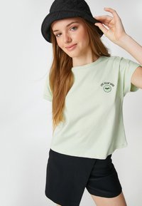 Camiseta de algodón verde claro con mangas cortas, que presenta un gráfico bordado de gafas de sol y el texto "Me siento bien" en el pecho. Sombrero de cubo negro.