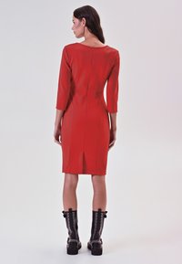 Rotes Kleid mit Dreiviertelärmel, taillierter Silhouette, Mittelnähten und einem Reißverschluss im Rücken. Kombiniert mit kniehohen schwarzen Stiefeln mit Nieten.
