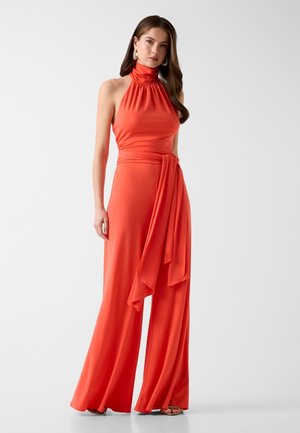 Donna che indossa una tuta arancione brillante senza maniche con collo alla coreana, pantaloni a gamba larga e una lunga fascia di tessuto in vita, in piedi contro uno sfondo bianco.