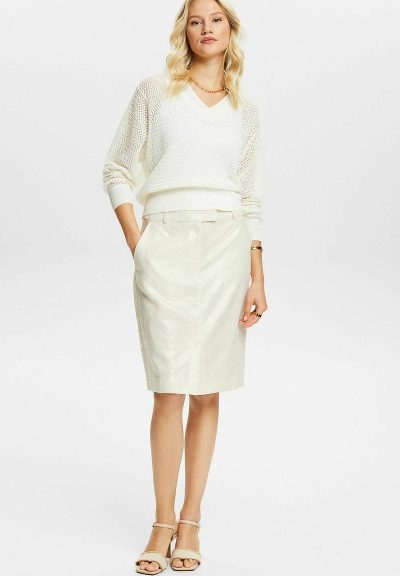 Femme blonde portant un sweater en maille blanc à manches longues, une jupe crème longueur genou, un collier en or, des bagues, un bracelet et des sandales beiges, se tenant avec les mains dans les poches.