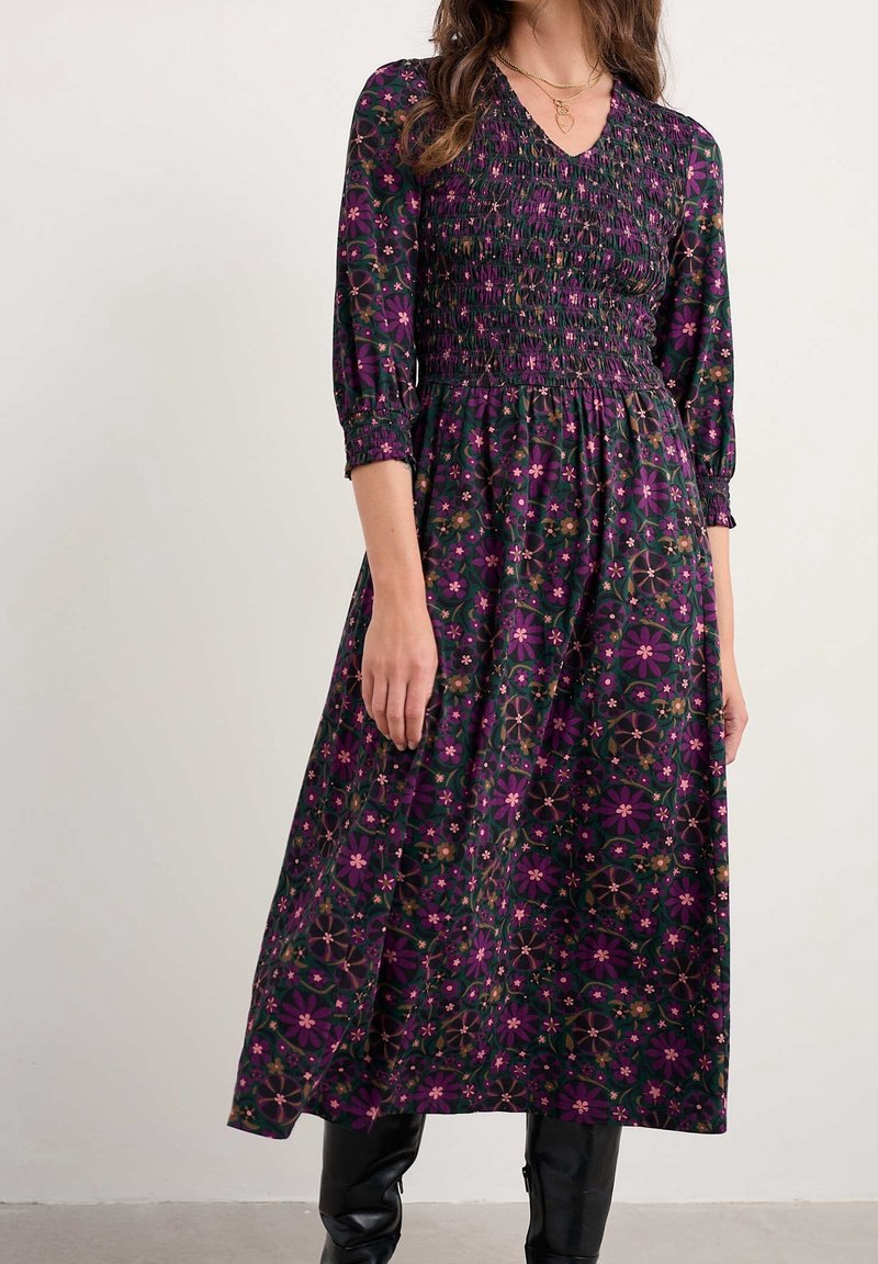 Femme portant une robe longue sombre à manches longues avec des motifs floraux violets et des bottes noires, debout devant un fond clair uni.