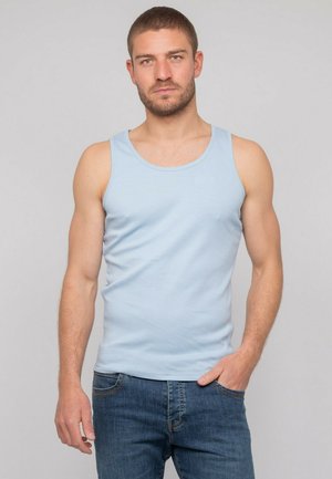 Homme aux cheveux bruns courts, portant un débardeur bleu clair et un jean bleu, debout devant un fond gris.