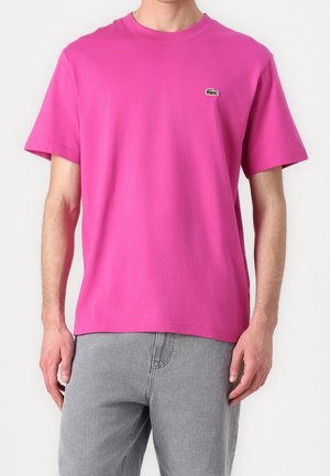 Jasnoróżowa bawełniana koszulka z krótkim rękawem, okrągłym dekoltem i małym zielonym haftowanym logo po lewej stronie klatki piersiowej, zestawiona z szarymi dżinsami.