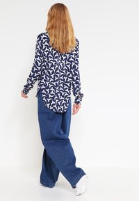 Camicia navy con un motivo di delfini bianchi, polsini con bottoni, spacchi laterali, abbinata a pantaloni di denim a gamba larga e sneakers bianche.