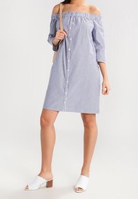 Randig off-shoulder klänning i blått och vitt, knälång, med trekvartslånga ärmar och knäppning fram. Bärs med vita mules.