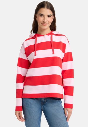 Young woman wearing a red and pink wide striped hoodie and blue jeans, standing with hands relaxed by sides.