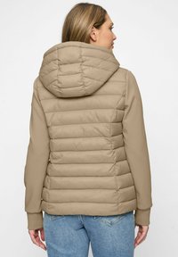 Frau, die mit dem Rücken zur Kamera steht, trägt eine beigefarbene, gesteppte Kapuzenjacke mit Fleece-Ärmeln und blaue Jeans vor einem einfachen, hellen Hintergrund.