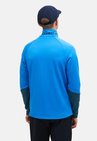 Camicia a maniche lunghe blu con colletto alto, caratterizzata da dettagli verde scuro sulle maniche e una texture liscia. Marchio "Perform" sul colletto.