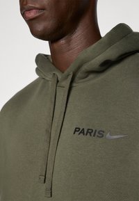 Felpa verde oliva con cordini, testo nero "PARIS" e logo Nike argento. Texture del tessuto liscia, design casual, angolazione ravvicinata.