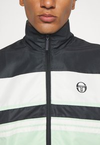 Juoda ir balta zip-up striukė su žaliais akcentais, su aukštu apykakle ir logotipu ant krūtinės, pagaminta iš tekstūruotos medžiagos.