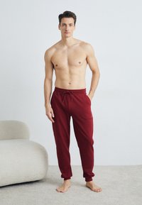 Burgunderfarbene Jogginghosen mit einem Kordelzugbund, schmal zulaufenden Beinen und dezentem Logodetail, vor einem einfarbigen Hintergrund und einer weich strukturierten Oberfläche.