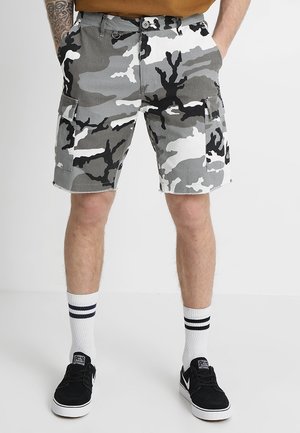 Homme portant un short cargo camouflage gris, blanc et noir, des chaussettes blanches à rayures noires et des baskets basses noires.