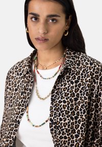 Leopard print shirt with rolled sleeves over a white top, adorned with multiple layered necklaces featuring colorful beads and gold accents.
