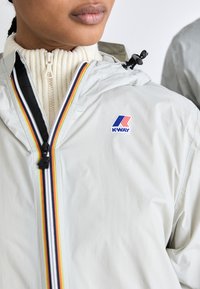 Light gray K-Way jacket with a hood, features a colorful vertical stripe along the zipper and a prominent logo on the chest. Textured fabric.