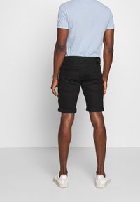 Pantalones cortos negros con dobladillos enrollados, hechos de denim, combinados con una camiseta polo azul claro y zapatillas blancas; se muestra desde una vista trasera.