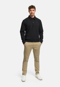 Czarna bluza polo z kołnierzykiem i dwoma guzikami, noszona na jasnobeżowych spodniach; model stoi z rękoma w kieszeniach, na stopach białe sportowe buty.
