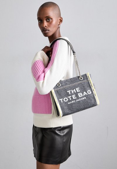 Sac en toile gris avec texte blanc, accents jaunes et poignées en chaîne métallique, présenté à côté d'un mannequin portant un pull rose et blanc.