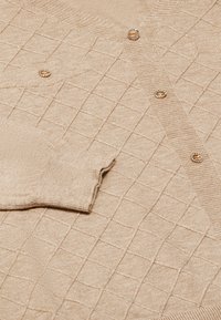 Cardigan beige con un motivo a griglia texture, dotato di bottoni color oro e polsini a costine. Realizzato in materiale morbido e leggero.