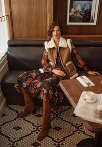 Gilet marrone con colletto in pelliccia su un vestito nero con stampa floreale. Stivali al ginocchio in suede marrone. Tavolo di legno con posate e una tazza di caffè.